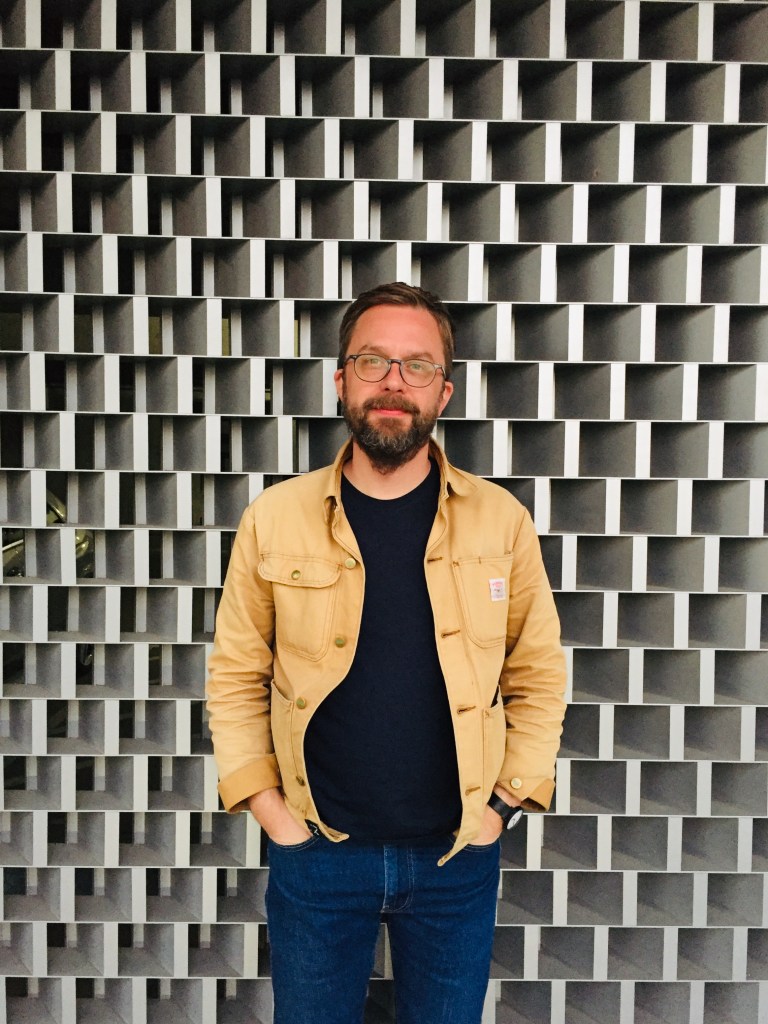 A man with glasses and a beard stands against a textured grey wall, wearing a beige jacket and dark shirt, with a relaxed expression.
